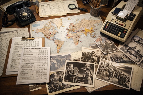 Overhead view of a 1950s film studio office desk with budget ledgers, box office reports, international distribution maps with pins, promotional stills from biblical epics, an adding machine, and a rotary phone.