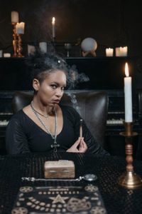 Woman holding incense with Ouija board on table and crystal ball on shelf