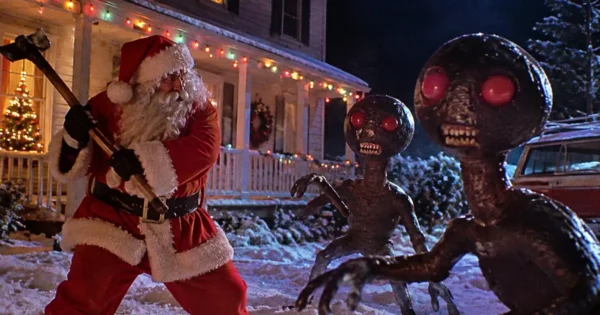Feeders 2: Slay Bells Review hero showing Santa Claus battling two red-eyed aliens outside a snow-covered house decorated for Christmas.