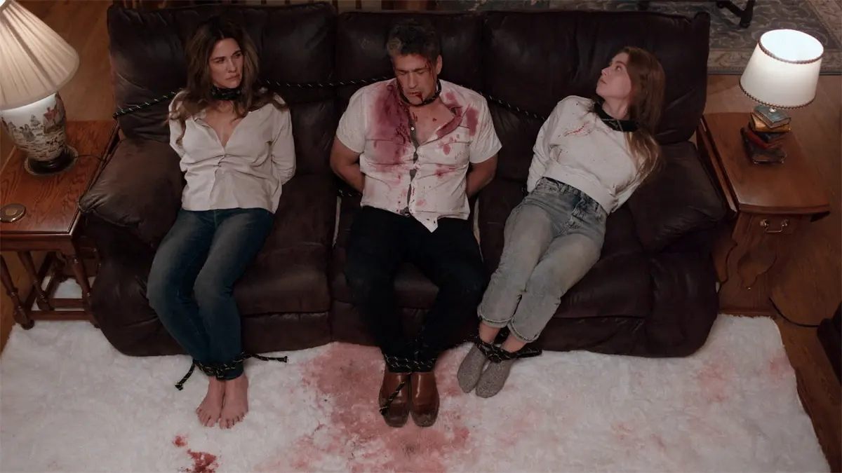 Malicious 2023 Review showing a terrified family tied up on a couch in a tense scene from the horror thriller.